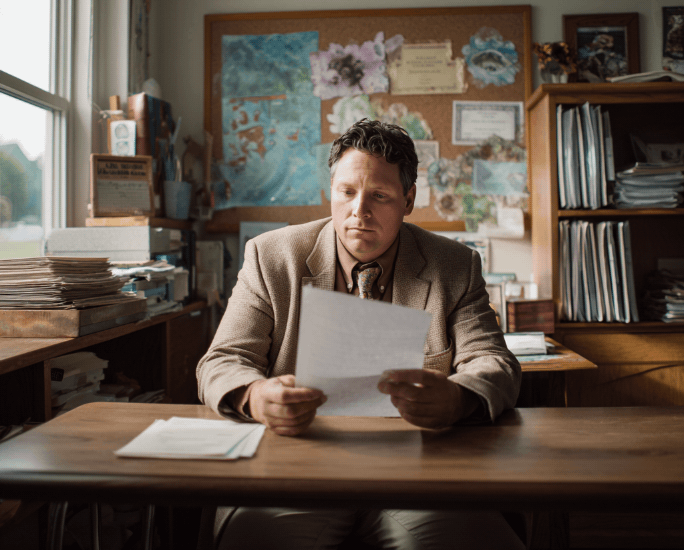 Male school principal at a wooden desk reading a report on educator injuries and staff retention.